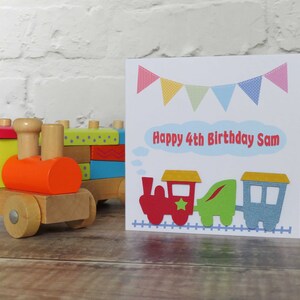 May include: A square greeting card featuring a colorful train illustration, bunting flags, and the text Happy 4th Birthday Sam, displayed next to a wooden toy train with a red engine, green middle car, and blue caboose on a track.