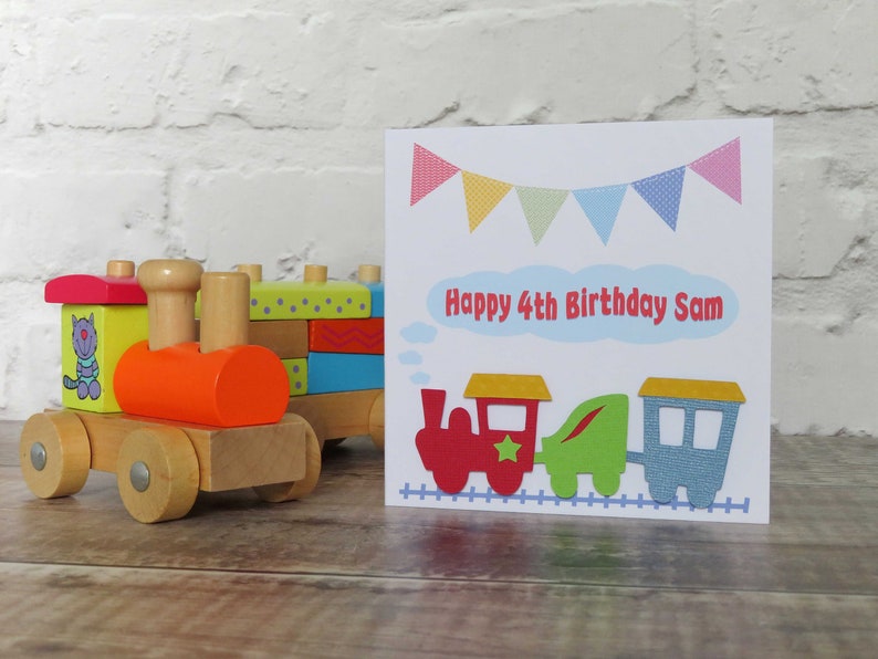 May include: A square greeting card featuring a colorful train illustration, bunting flags, and the text Happy 4th Birthday Sam, displayed next to a wooden toy train with a red engine, green middle car, and blue caboose on a track.