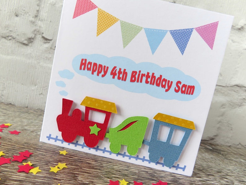 May include: A white birthday card featuring a colorful bunting banner at the top and a light blue cloud shape with the red text Happy 4th Birthday Sam. Below, a three-piece train in red, green, and blue sits on a blue train track graphic.