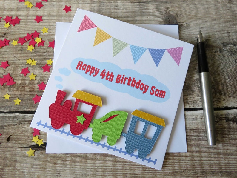 May include: A square white greeting card featuring a colorful train illustration with a red engine, green carriage, and blue caboose on a light blue track. Above the train, a light blue cloud contains the red text Happy 4th Birthday Sam.