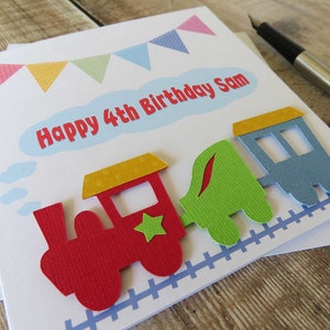 May include: A white birthday greeting card featuring a colorful paper train cutout with a red engine, green carriage, and blue caboose. The card displays the text Happy 4th Birthday Sam in red letters inside a light blue cloud shape, with bunting above.