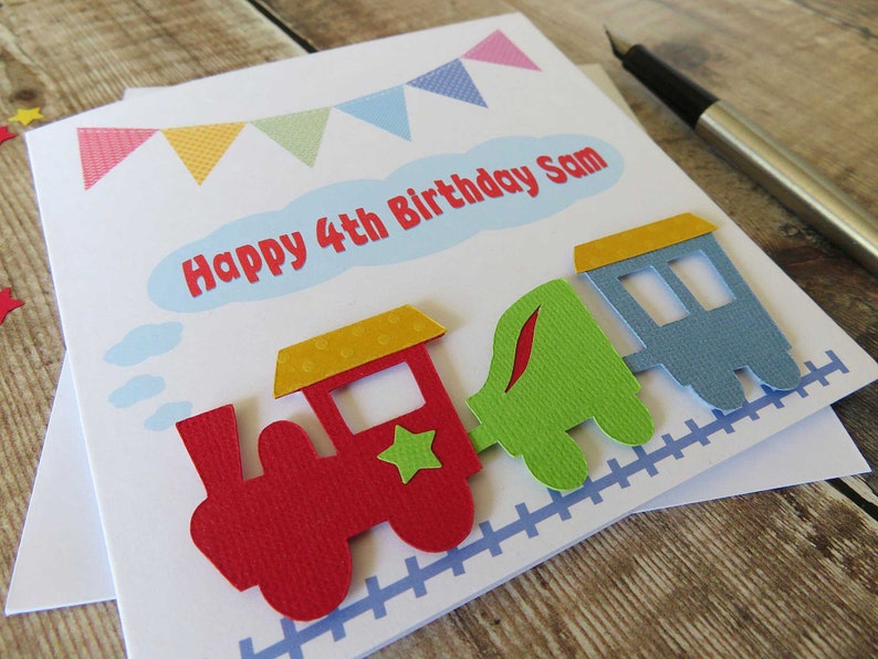 May include: A white birthday greeting card featuring a colorful paper train cutout with a red engine, green carriage, and blue caboose. The card displays the text Happy 4th Birthday Sam in red letters inside a light blue cloud shape, with bunting above.