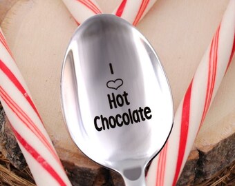 May include: A silver spoon with the words "I love Hot Chocolate" engraved on it. The spoon is resting on a wooden slice with a red and white candy cane wrapped around it. A mug of hot chocolate with marshmallows is in the background.