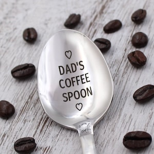 May include: A silver coffee spoon with the words "Dad's Coffee Spoon" engraved on the bowl. The spoon is surrounded by coffee beans on a white wooden surface.