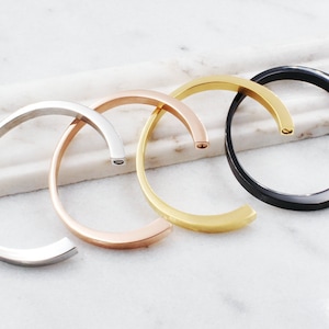 May include: Four simple bangle bracelets in silver, rose gold, gold, and black metal. The bracelets are stacked on a white marble surface.
