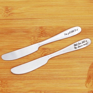 May include: Two silver butter knives with engraved text. The top knife reads "You just fold it in!" and the bottom knife reads "What does fold in the cheese mean?"