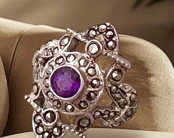 Puede incluir: Un anillo de plata con una piedra preciosa púrpura rodeada de un delicado y complejo diseño de pequeñas piedras brillantes.