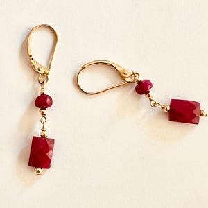 Könnte beinhalten: Ein Paar goldfarbene Ohrringe mit Klappbügelverschluss. Jeder Ohrring hat einen kleinen, runden, roten Edelstein, eine goldene Perle und einen größeren, rechteckigen, facettierten roten Edelstein. Die Ohrringe werden vor einem hellen Hintergrund präsentiert.