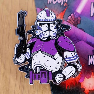 May include: A purple and white enamel pin depicting a clone trooper with a blaster. The pin has a black outline and a cosmic background. The trooper's armour is detailed with purple accents and is a collectible item.