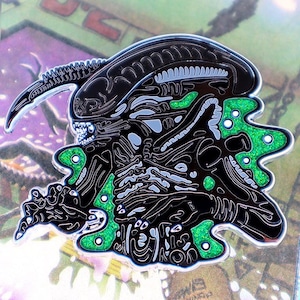May include: A black and silver enamel pin depicting an Alien creature with green glitter accents. The pin showcases detailed line work, highlighting the creature's distinctive features and overall design.