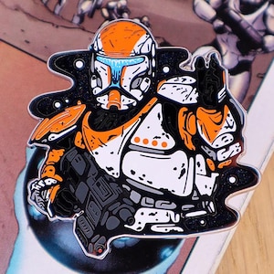 May include: Enamel pin depicting a soldier in a helmet and armour. The pin has a black outline with orange, white, grey, and blue details. The soldier is holding a weapon. The pin is on a comic book page.