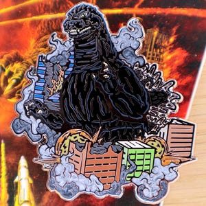 May include: A black Godzilla enamel pin with a detailed design. The pin features Godzilla amidst a cityscape, with buildings and smoke. The background shows an orange and red fiery scene. The pin is a collectible item.