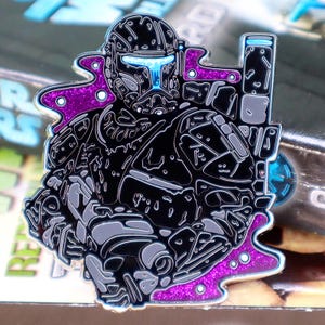 Puede incluir: Un pin de esmalte detallado que representa una figura blindada negra y gris con una visera y acentos azules brillantes. El pin tiene un borde de purpurina morada y es probablemente un artículo de colección.