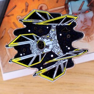 May include: A detailed enamel pin featuring a TIE fighter spacecraft against a black starry background. The pin has yellow and white accents, with a silver-colored border. The design is inspired by a science fiction theme.