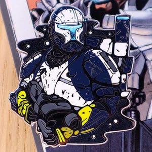 May include: An enamel pin depicting a futuristic soldier in dark blue and white armour. The pin has a black outline with yellow and grey accents, and a weapon is visible. The design is reminiscent of science fiction or video game themes.