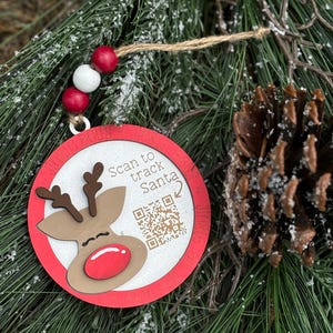 May include: A round Christmas ornament with a red border and a reindeer design. The ornament has the text "Scan to track Santa" and a QR code. It is strung with a twine and red and white beads. The ornament is set against a backdrop of pine needles and pine cones.