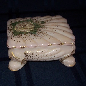 May include: A pink glass trinket box with a shell design and gold accents. The lid features a painted flower with green leaves. The box sits on four feet shaped like fish.