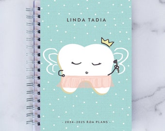 May include: A teal blue planner with white polka dots and a cartoon tooth fairy illustration. The planner has a clear plastic cover and a silver spiral binding. The text "LINDA TADIA" and ". 2024-2025 RDA PLANS." are printed on the cover.