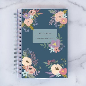 May include: A blue planner with a floral design and the text "KATIE WEST - 2024-2025 RDA PLANS -" on the cover.