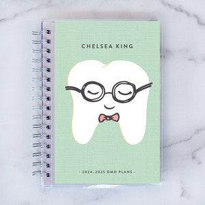 May include: A spiral-bound planner with a clear plastic cover. The cover is light green and features a cartoon tooth wearing glasses and a bow tie. The text "CHELSEA KING" and "2024-2025 DMD PLANS" are printed on the cover.