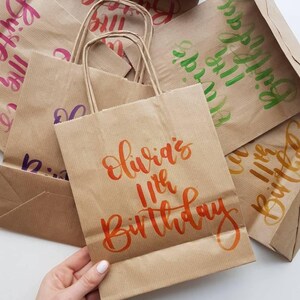 May include: Brown paper gift bags with handwritten calligraphy in various colors. The bags are decorated with the words "Happy Birthday" and "Olivia's 11th Birthday".