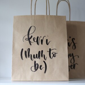 May include: Two brown paper gift bags with black handwritten text. The bag on the left says "Kerri (Mum to be)" and the bag on the right says "This is my forever".