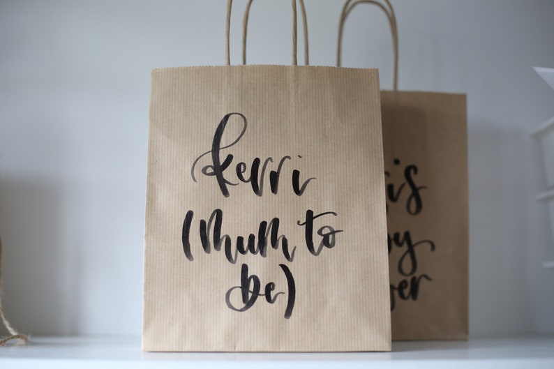 May include: Two brown paper gift bags with black handwritten text. The bag on the left says "Kerri (Mum to be)" and the bag on the right says "This is my forever".