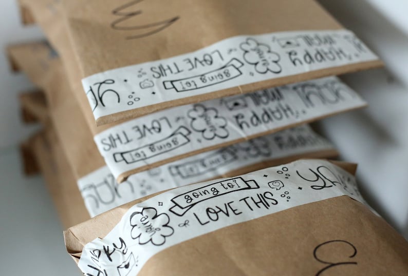 Puede incluir: Una pila de paquetes de papel marr&oacute;n con cinta blanca y escritura con marcador negro. La cinta tiene un dise&ntilde;o de nube con las palabras "LOVE THIS" y "going to!" escritas en ella.