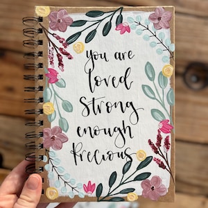 Puede incluir: Un cuaderno con espiral con un diseño floral pintado. La portada presenta las palabras "you are loved strong enough precious" en escritura negra, rodeadas de flores y hojas pintadas en rosa, amarillo y verde.