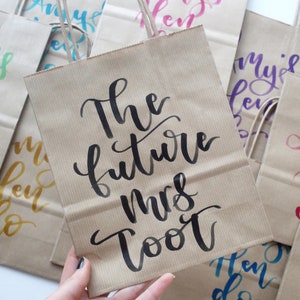 May include: Brown paper gift bags with black calligraphy writing. The text on the front of one bag reads "The future Mrs. [Last Name]".