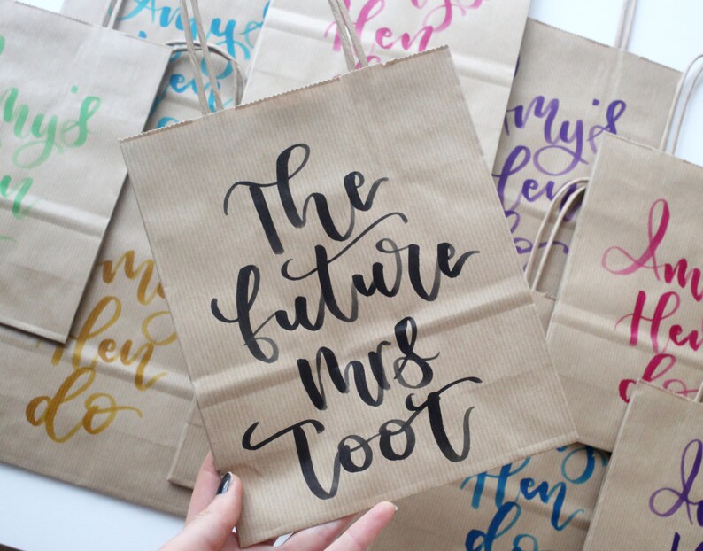 May include: Brown paper gift bags with black calligraphy writing. The text on the front of one bag reads "The future Mrs. [Last Name]".