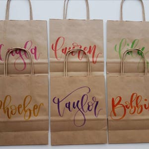 May include: Six brown paper gift bags with handles. Each bag has a different name written on it in colorful calligraphy: Cayla, Lauren, Lily, Phoebe, Taylor, and Bobbie.