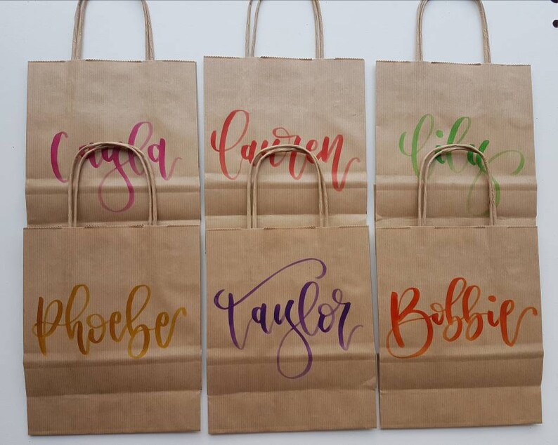 May include: Six brown paper gift bags with handles. Each bag has a different name written on it in colorful calligraphy: Cayla, Lauren, Lily, Phoebe, Taylor, and Bobbie.