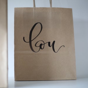 May include: Two brown paper shopping bags with handles. The bag on the right has the word "Lou" written in black cursive on the front.