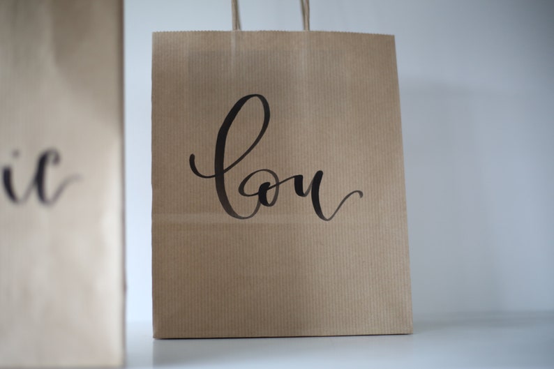 May include: Two brown paper shopping bags with handles. The bag on the right has the word "Lou" written in black cursive on the front.