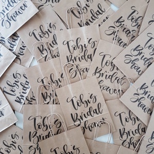 May include: A collection of brown paper gift bags with black calligraphy that reads "Tobi's Bridal Shower".