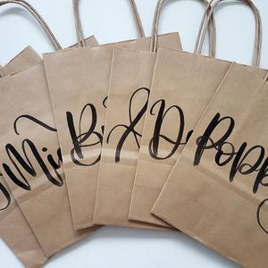 May include: Six brown paper gift bags with black calligraphy writing on the front. The bags are labeled "Mimi", "B", "D", "Poppy", and two other names that are not visible in the image.