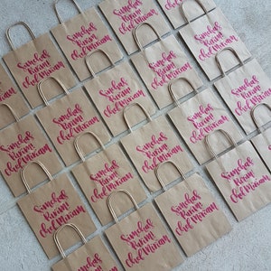 May include: A collection of brown paper bags with handles, each decorated with the text "Simchas Purim! Chef Miriam" in pink lettering.