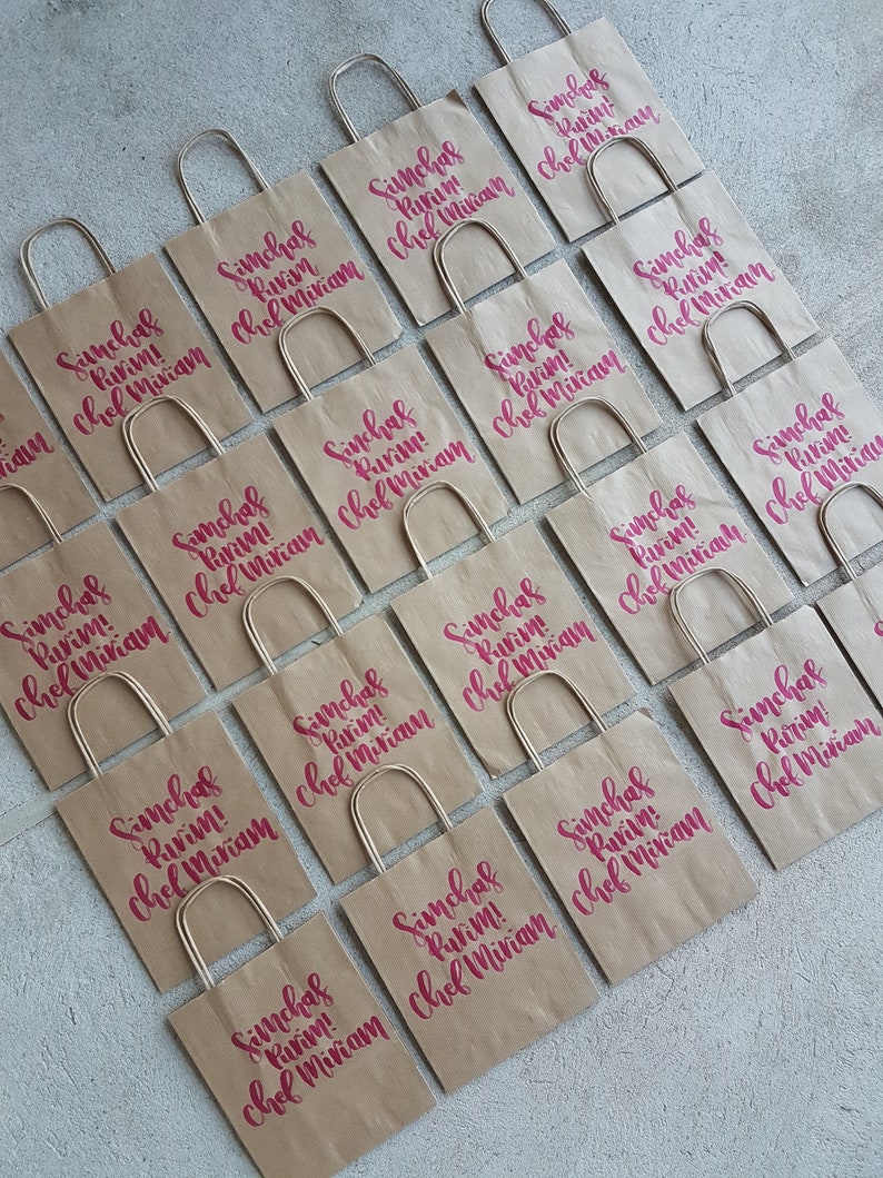 May include: A collection of brown paper bags with handles, each decorated with the text "Simchas Purim! Chef Miriam" in pink lettering.