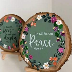 Puede incluir: Dos placas redondas de madera con coronas de flores pintadas y citas inspiradoras. Una dice "Above all Love each other deeply", la otra "He will be our Peace - Micah 5:5". Las placas están sobre pequeños caballetes de madera.