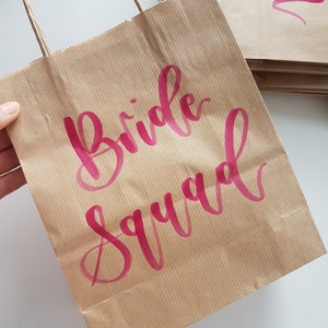 May include: Brown paper gift bag with a pink inscription that reads "Bride Squad".