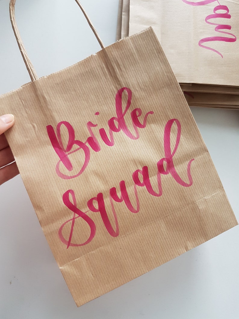 May include: Brown paper gift bag with a pink inscription that reads "Bride Squad".