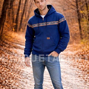 May include: A navy blue hooded zip-up jacket with a patterned design across the chest and shoulders. The jacket has a small rectangular patch on the front and is paired with blue jeans. The background shows a path through a forest.
