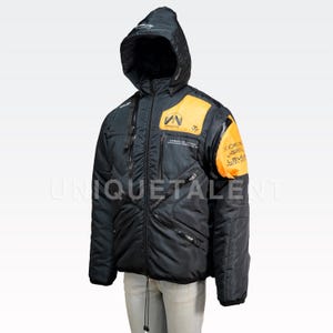 May include: Black hooded jacket with orange accents on the shoulders and chest. The jacket has multiple zippered pockets and a drawstring at the waist. Text is visible on the chest and sleeve. The jacket is designed for cold weather.
