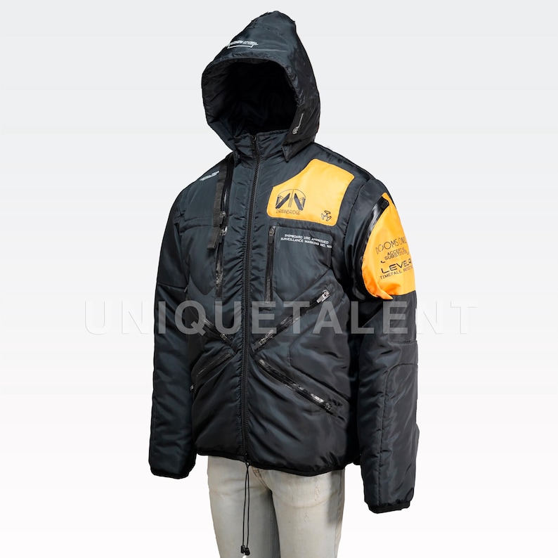 May include: Black hooded jacket with orange accents on the shoulders and chest. The jacket has multiple zippered pockets and a drawstring at the waist. Text is visible on the chest and sleeve. The jacket is designed for cold weather.