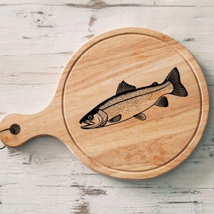 May include: Round wooden cutting board with a handle, featuring a detailed black illustration of a fish. The board has a natural wood grain finish and a small hole in the handle for hanging.