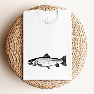 May include: White t-shirt featuring a black and white illustration of a fish. The fish is a detailed drawing with scales and fins. The shirt is displayed on a woven, circular surface.