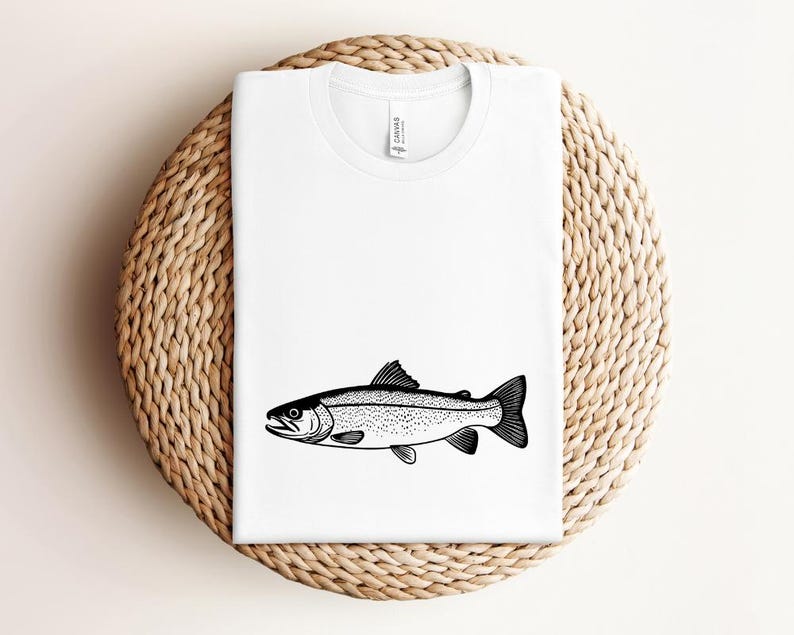 May include: White t-shirt featuring a black and white illustration of a fish. The fish is a detailed drawing with scales and fins. The shirt is displayed on a woven, circular surface.