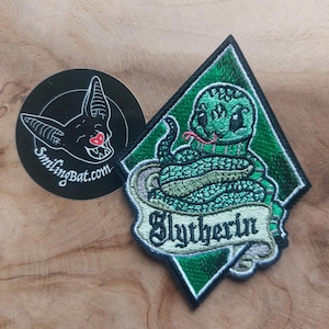 Op de afbeelding: Een groen en zwart geborduurde patch met een cartoon slang met het woord "Slytherin" op een banner. Een zwarte ronde sticker met een lachend vleermuislogo staat naast de patch. De patch is ruitvormig.
