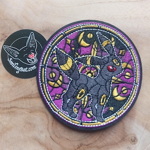 Puede incluir: Parche bordado con un diseño de Umbreon negro dentro de un marco de estilo vidriera. El parche tiene una combinación de colores púrpura y dorado, con un borde negro. También hay una pegatina circular con un logotipo de murciélago y el texto "smilingbat.com".